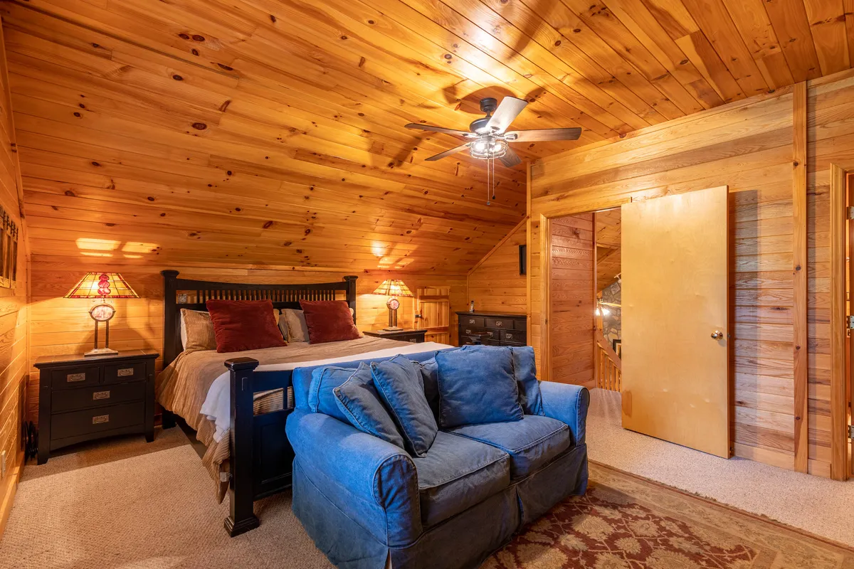 A cozy loft or den area featuring a bed adorned with decorative pillows, a soft blue sofa, a wooden ceiling, and bedside lamps. The relaxed setting is ideal for lounging, reading, or accommodating extra guests.
