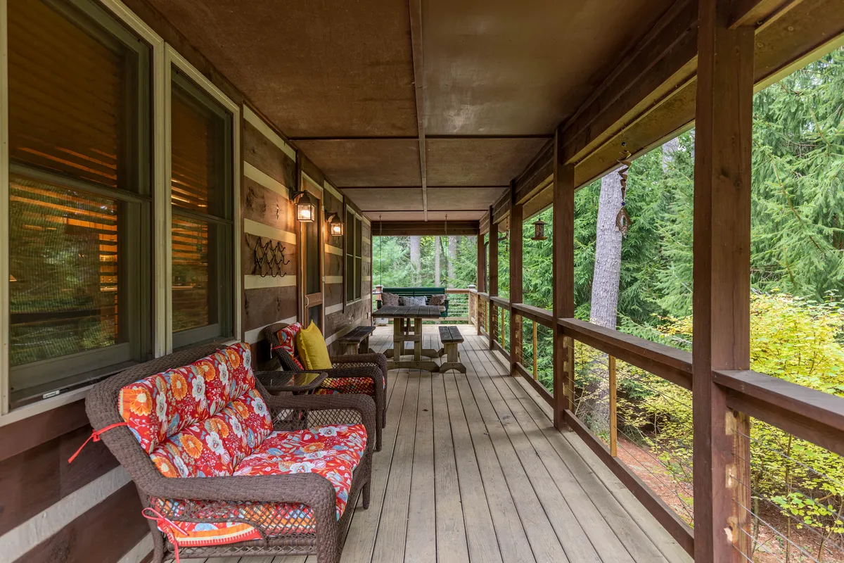 A cozy dining area on the deck with colorful patterned cushions, a table for outdoor meals or games, and a view of dense greenery beyond the wooden railing. The relaxed setup is perfect for gathering with friends or enjoying a quiet meal.