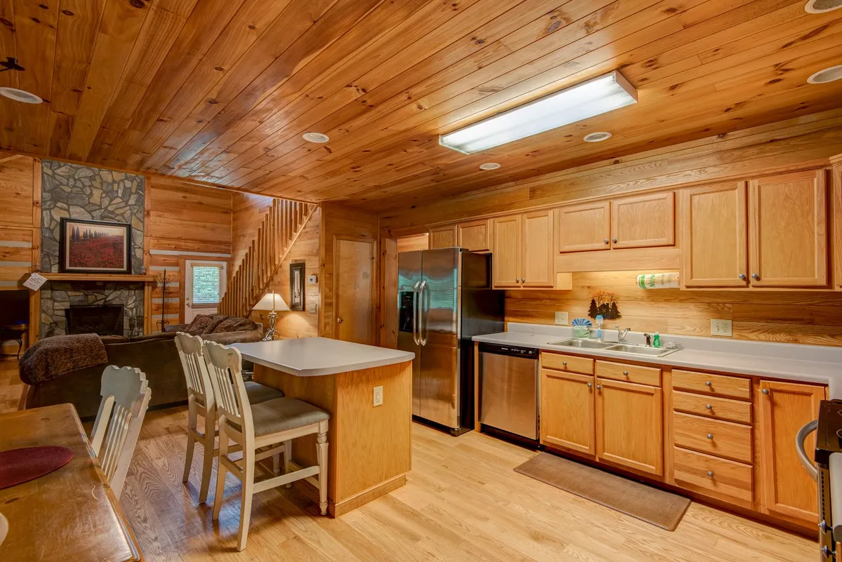 This image features an inviting, open kitchen area with a kitchen island, stainless steel appliances, and wooden cabinetry. The backdrop includes a stone fireplace, helping blend the kitchen and living room spaces for a homey, all-season feel. The cozy textures and abundant counter space suggest it is ideal for meal prep, entertaining, or unwinding after outdoor adventures.