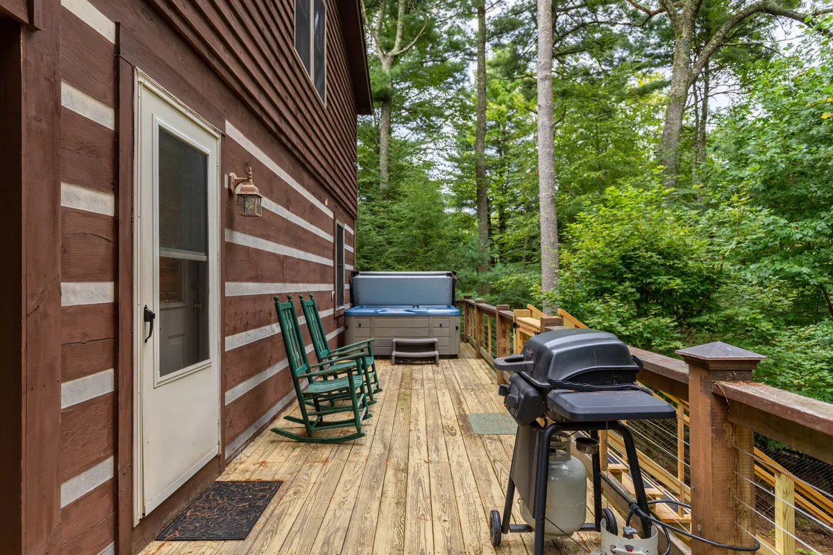 This image captures a spacious, wooden deck featuring rocking chairs, a bubbling hot tub, and a gas grill. The deck is bordered by a rustic railing and set against a lush, wooded backdrop, with dappled sunlight filtering through the trees.