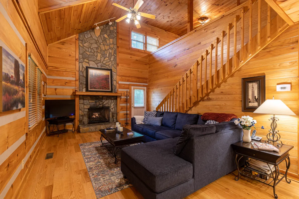 This image highlights the main living area, featuring a striking stone fireplace as the focal point. The cozy sofa and wooden staircase contribute to the welcoming, open layout, while artwork and rustic decor create a cabin ambiance. The presence of the fireplace and plush seating perfectly capture the communal, relaxing vibe described in the section.