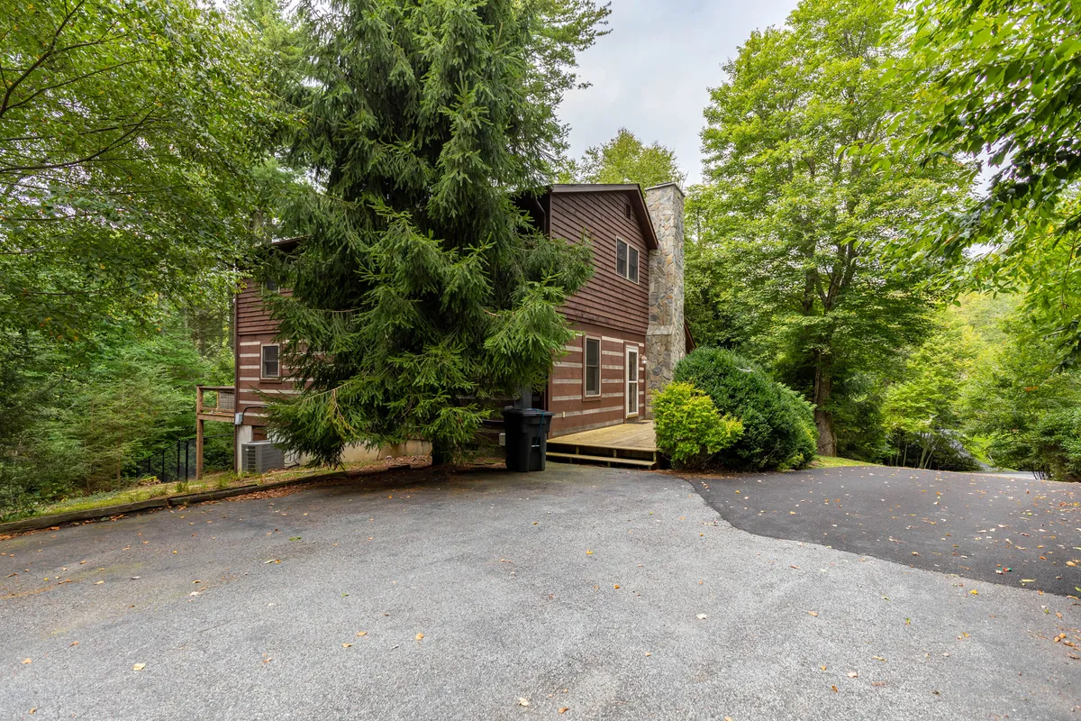 This image shows the exterior of a rustic wooden house surrounded by a large evergreen tree and landscaped shrubs. The home sits in a serene, nature-filled setting that invites outdoor relaxation and exploration. The landscaping and large deck area make it ideal for morning coffee, evening bonfires, or simply soaking in the mountain air.