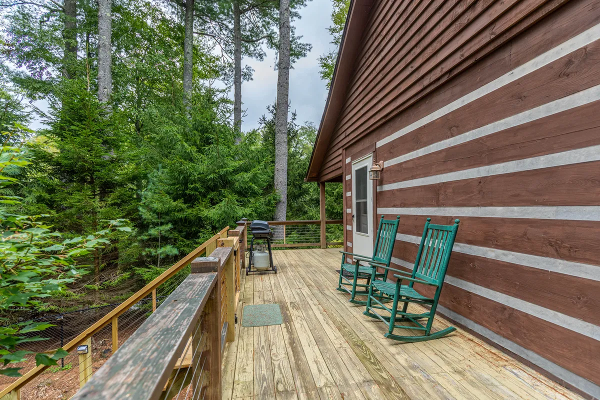 A tranquil wooden deck furnished with rocking chairs and a grill, surrounded by lush greenery. The scene suggests leisurely afternoons, outdoor gatherings, or peaceful moments listening to nature while enjoying a cup of tea or a fresh barbecue meal.