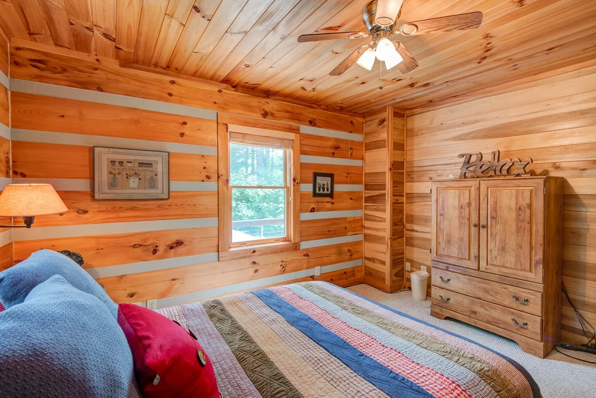 A serene queen bedroom with wooden walls, a colorful quilt on the bed, a window offering views to nature, a nightstand with a lamp, and a wardrobe with a 'Relax' sign. The soft lighting and peaceful atmosphere evoke comfort and restfulness.