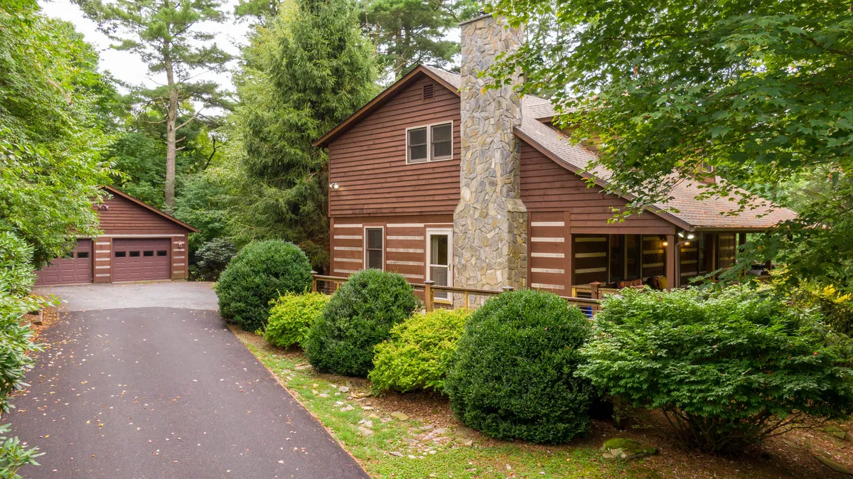 The image showcases a charming two-story log cabin with a striking stone chimney and rustic wooden siding, set amid a beautifully landscaped front yard. The welcoming front porch and spacious yard invite guests to relax and enjoy the serene mountain surroundings. The garage and stone pathways add to the sense of a well-appointed, inviting retreat nestled in nature.