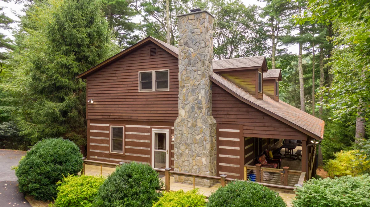 A cozy exterior view with a stone chimney, wooden siding, and landscaped greenery sets the scene for family gatherings, barbecues, and outdoor relaxation. The space is welcoming, with a porch ideal for stargazing or morning coffee surrounded by nature.